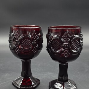 May include: Two ruby red stemmed wine glasses. The glasses have a detailed, textured pattern around the bowl and a faceted base. The stem is a solid, dark red color. The glasses are displayed against a dark background.