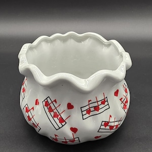 Può includere: Ciotola in ceramica bianca con bordo a balze. La ciotola è decorata con cuori rossi e note musicali. Il disegno si ripete intorno all'esterno della ciotola. La ciotola viene probabilmente utilizzata per scopi decorativi.