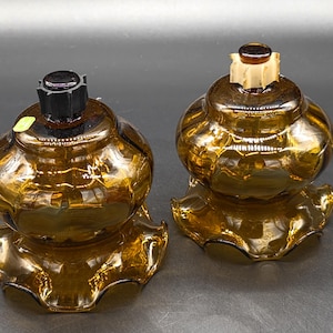 May include: Two amber-colored glass candle holders with a ruffled base and a round body. One has a black top, the other has a tan top. The candle holders are on a dark gray surface.