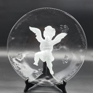 May include: Clear glass plate with an etched design of a cherub with wings, surrounded by roses and butterflies. The plate is displayed on a black stand, showcasing the intricate details of the artwork. The plate is round.