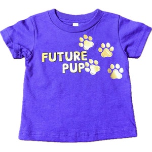 Puede incluir: Camiseta de manga corta de color morado con texto en purpurina dorada que dice "FUTURE PUP" y huellas de pata en purpurina dorada.