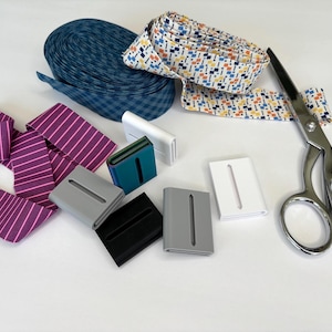 May include: An assortment of sewing supplies, including rolls of patterned fabric, fabric clips in various colors, and a pair of silver scissors. The fabric includes blue gingham, purple stripes, and a colorful geometric print.