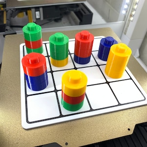 May include: A 3D printed game board with a grid of nine squares.  Each square has a stack of colorful cylinders, red, blue, green, and yellow.