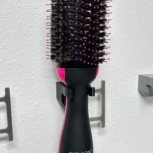 May include: A black and pink hot air brush with a round brush head. The brush is mounted on a white wall with a gray metal hook.