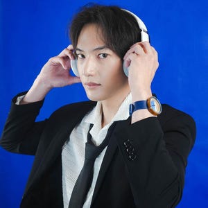 May include: A man wearing a black suit jacket over a white shirt with a black tie. He is wearing a blue and brown wristwatch and white headphones over his ears.