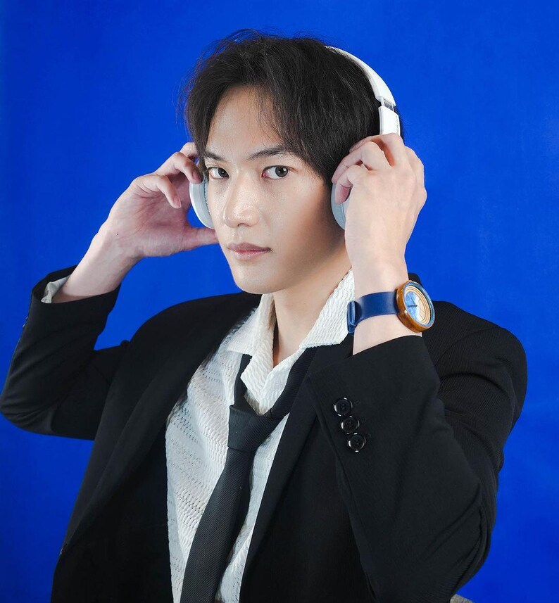 May include: A man wearing a black suit jacket over a white shirt with a black tie. He is wearing a blue and brown wristwatch and white headphones over his ears.