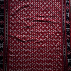 Könnte beinhalten: Ein rechteckiger, roter Stoff mit einem sich wiederholenden weißen Blumenmuster. Die Ränder weisen schwarz-weiße geometrische Designs auf. Der untere Rand hat einen Fransen. Dieser Stoff könnte als Schal oder Dekostoff verwendet werden.