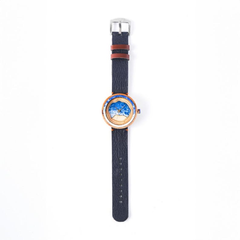 May include: A wooden wristwatch with a blue and white ocean wave design on the face. The watch has a black leather band with a brown leather accent.
