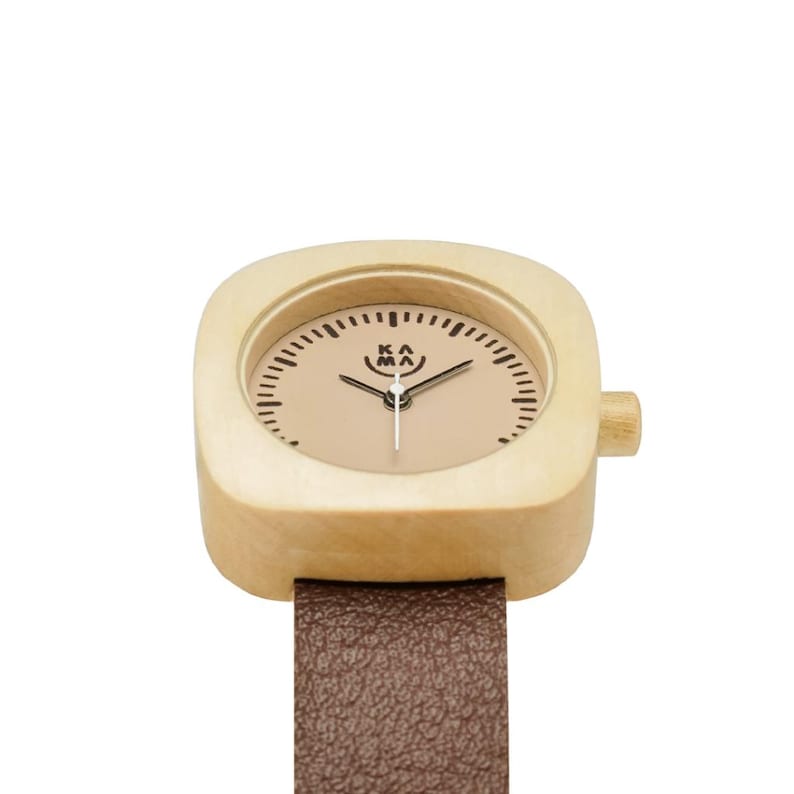 May include: A wooden wristwatch with a square face and a brown leather band. The watch face is tan with black markings and the text "KA MA". The watch has a wooden crown on the side.