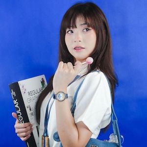 May include: A young person with long dark hair is wearing a white shirt and a blue denim overall dress. They are holding a book titled "Regular Graphic Design" and a lollipop. They are also wearing a watch with a gold face and blue band.