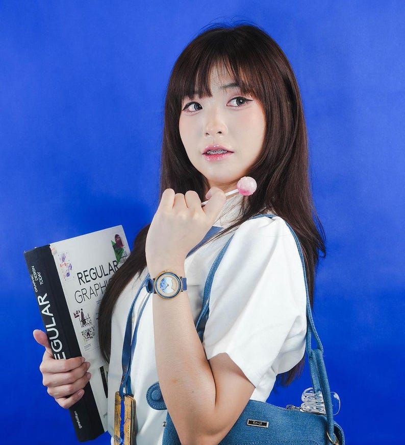 May include: A young person with long dark hair is wearing a white shirt and a blue denim overall dress. They are holding a book titled "Regular Graphic Design" and a lollipop. They are also wearing a watch with a gold face and blue band.