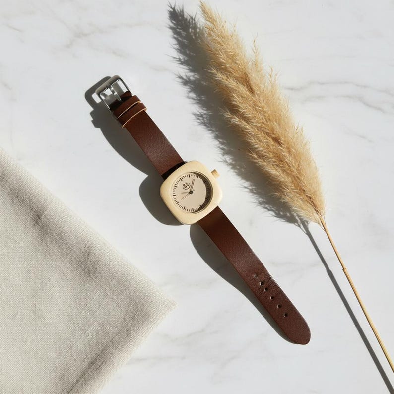 May include: A wristwatch with a square, cream-colored face and a brown leather band. The watch has a silver buckle and a small crown. The watch is lying on a white marble surface next to a beige cloth and a dried plant.