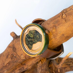 May include: A wooden wristwatch with a green and brown face and a brown leather band. The watch is sitting on a piece of driftwood.