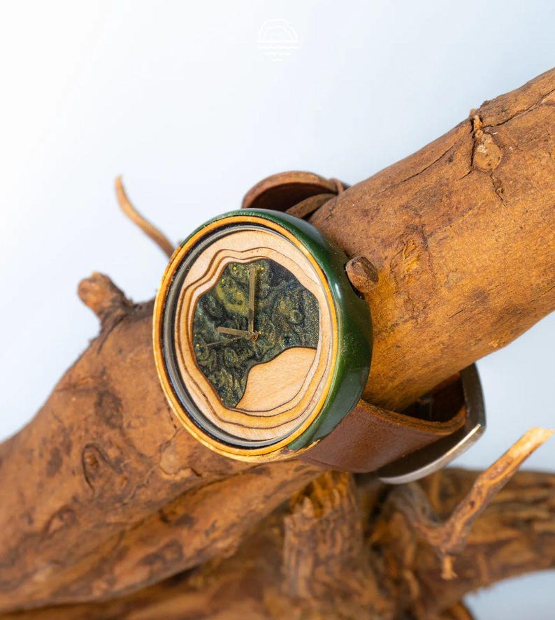 May include: A wooden wristwatch with a green and brown face and a brown leather band. The watch is sitting on a piece of driftwood.