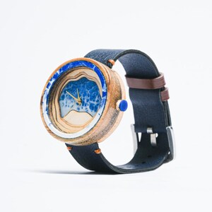 May include: A wooden wristwatch with a blue and white wave design on the face. The watch has a dark blue leather band with a brown leather lining.
