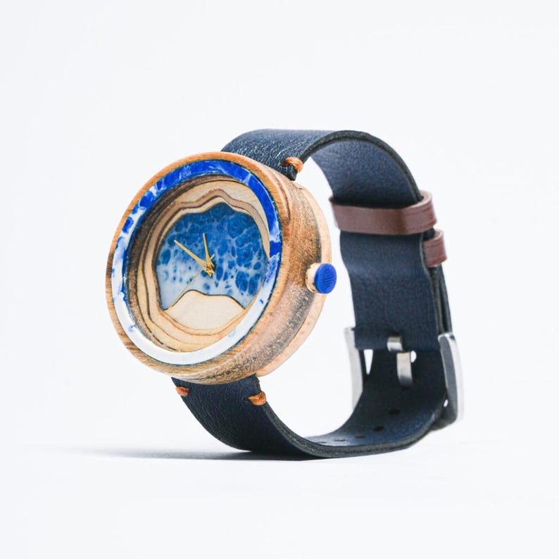 May include: A wooden wristwatch with a blue and white wave design on the face. The watch has a dark blue leather band with a brown leather lining.
