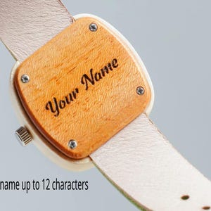 May include: Wooden watch with a light-colored leather strap. The watch face is a light brown wood with the engraved text "Your Name". The watch face is square with rounded corners and is secured with four small screws.