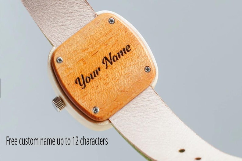May include: Wooden watch with a light-colored leather strap. The watch face is a light brown wood with the engraved text "Your Name". The watch face is square with rounded corners and is secured with four small screws.