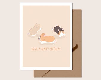 May include: A greeting card with three cartoon corgi dogs in various colors on a light peach background. The text "HAVE A FRAPPY BIRTHDAY" is printed below the dogs. The card is inside a white border and a brown envelope.