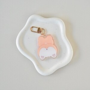 Op de afbeelding: Een heldere acryl sleutelhanger met een cartoon corgi kont ontwerp in tinten perzik, wit en roze. De sleutelhanger heeft een goudkleurige sluiting. Het corgi kont ontwerp bevat pootafdrukken en een klein zwart kruis.