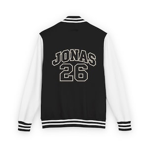Peut inclure: Une veste universitaire noire avec des manches blanches et des bords-côtes rayés au col, aux poignets et à la taille. Le dos présente le texte JONAS et le numéro 26 dans une police de style universitaire blanche avec un fin contour noir.