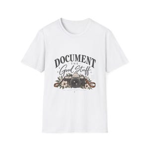 Puede incluir: Una camiseta blanca de manga corta con un gráfico de una cámara vintage rodeada de flores y hojas. El texto "DOCUMENT THE Good Stuff" aparece en el centro del pecho en una combinación de fuentes serif en negrita y cursiva.