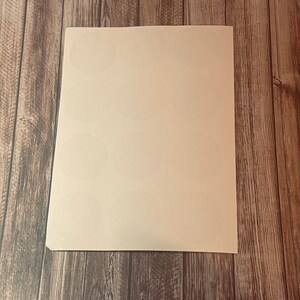 May include: A sheet of white, circular stickers on a white background. The stickers are arranged in rows and columns. The sheet is placed on a wooden surface with a natural wood grain pattern.