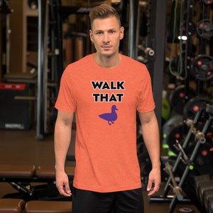 Puede incluir: Camiseta naranja de manga corta con el texto "WALK THAT" en letras negras y una silueta de pato morada debajo del texto.