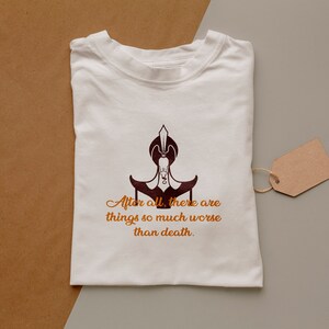 May include: White t-shirt with a brown graphic of a character from the movie Aladdin. The graphic is of the character Jafar, sitting on his throne. The text below the graphic reads "After all, there are things so much worse than death."