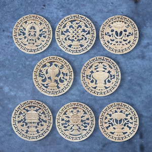 Puede incluir: Ocho adornos circulares de madera tallada. Cada adorno presenta un diseño diferente, como flores de loto y patrones geométricos. Los adornos de color marrón claro están dispuestos sobre una superficie azul. Cada adorno mide aproximadamente 10 cm de diámetro.