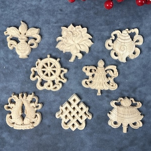 Puede incluir: Una colección de nueve adornos de madera tallada intrincadamente en varias formas, incluyendo una flor de loto, una rueda y un jarrón. Los adornos son de color marrón claro y están dispuestos sobre una superficie azul oscuro.