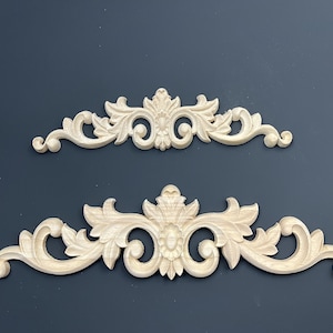 Può includere: Due applicazioni decorative in legno non rifinite, di colore chiaro, con un design simmetrico e ornato. Le applicazioni presentano un motivo floreale centrale con dettagli di foglie e volute. Una è più piccola, l'altra più grande.