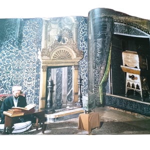 May include: A double-page spread from a book featuring a man in traditional clothing reading. The left page shows the man seated at a small table, with ornate wall patterns and architectural details. The right page displays a detailed interior with a decorative cabinet.