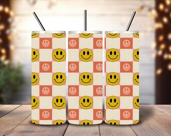 May include: Three stainless steel tumblers with black straws. The tumblers feature a repeating pattern of yellow smiley faces on a cream and coral checkerboard background. The tumblers are cylindrical and sit on a wooden surface.
