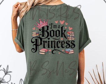 Puede incluir: Camiseta verde oliva con el texto "I'm a Book Princess" en fuente negra. El diseño incluye una corona rosa, corazones rosas y pilas de libros de varios colores. La camiseta tiene mangas enrolladas.