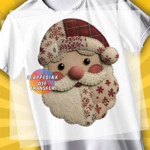 May include: White t-shirt featuring a Santa Claus face design. The Santa face is made of fabric with a patchwork design, including a red hat with snowflakes, a round pink nose, and a white beard. The text "SLAPPEDINK DTF TRANSFER" is also visible.