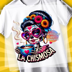 May include: White t-shirt featuring a colorful graphic of a sugar skull woman with heart-shaped glasses, holding a coffee cup. The design includes the text "LA CHISMOSA" and "SLAPPEDINK DTF TRANSFER". The woman has a floral crown and gold hoop earrings.