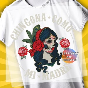 May include: White t-shirt featuring a colorful graphic of a sugar skull woman with long dark hair, a red headscarf, and red roses. The text "CHINGONA COMO MI MADRE" is printed above and below the graphic.
