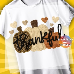 May include: A white t-shirt featuring a graphic of three pumpkins with a knit-like texture. The left pumpkin has a leopard print, the center is cream, and the right is orange. The word "thankful" is written in black script over the pumpkins.