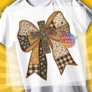 May include: White t-shirt featuring a large, colorful bow design with a cross at the center. The bow is made of various patterned fabrics, including floral, gingham, and solid colors. The text "SLAPPEDINK DTF TRANSFER" is visible.