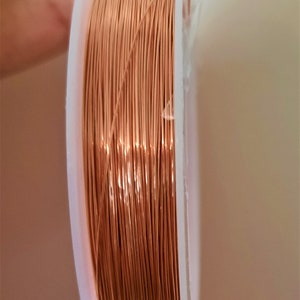 May include: A spool of copper-coloured wire, tightly wound on a white plastic spool. The wire is thin and shiny, reflecting light. Suitable for jewellery making or craft projects.