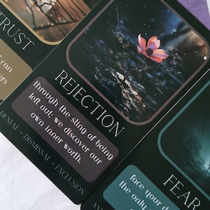 May include: A close-up of tarot cards with dark backgrounds and white text. One card reads "REJECTION" with the text "through the sting of being left out; we discover our own inner worth." Another card reads "FEAR".