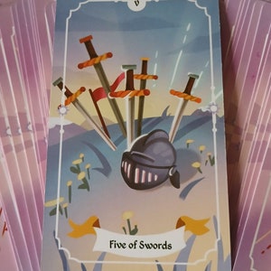 May include: A tarot card from a deck, featuring a helmet and five swords. The card is labeled "Five of Swords" and has a light blue background with a landscape design. The card is surrounded by other cards.