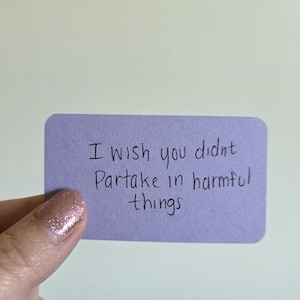 May include: A small, lavender-colored card with rounded corners. The handwritten text reads, "I wish you didn't Partake in harmful things." The card is held by a hand with glittery nail polish.