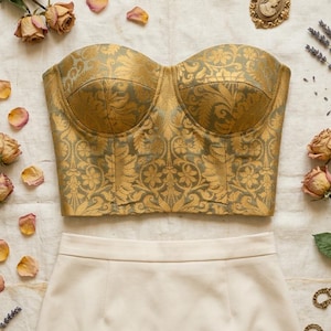 May include: A gold and green patterned bustier top paired with a cream-colored skirt and black heels. The ensemble is accessorized with a vintage pocket watch, cameo brooch, and dried roses, creating a vintage aesthetic.