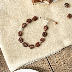 Könnte beinhalten: Ein silbernes Armband mit braunen Kaffeekern-Anhängern. Das Armband liegt auf einem beigefarbenen Handtuch mit Schokoladenquadraten im Hintergrund.