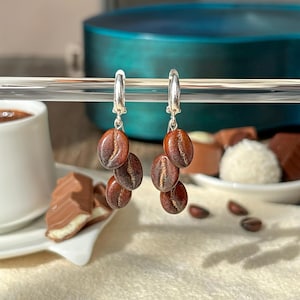 Könnte beinhalten: Silberne Creolen mit drei baumelnden Kaffeebohnen-Anhängern. Die Anhänger sind braun mit einem hellbraunen Streifen in der Mitte.
