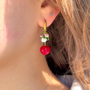 以下が含まれることがあります： 赤いチェリーチャームと白とピンクの花のアクセントが付いたゴールドのフープピアス