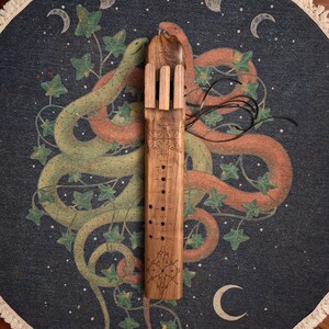 May include: A wooden flute with intricate carvings and a series of holes along its length. The flute is decorated with a geometric pattern and is resting on a fabric background with a snake design.