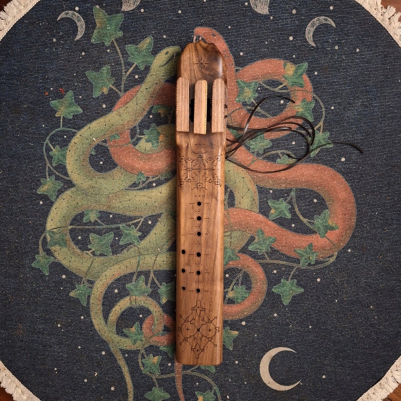 May include: A wooden flute with intricate carvings and a series of holes along its length. The flute is decorated with a geometric pattern and is resting on a fabric background with a snake design.